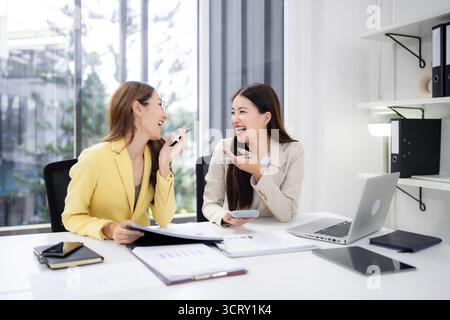 Two joyful adult Asian women engaged in productive conversation an office setting. They discuss business ideas enthusiastically. Happy colleagues coll Stock Photo