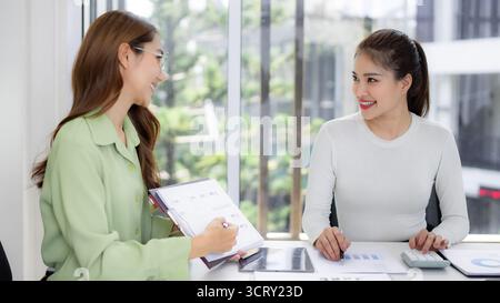 Adult Asian female professionals actively engage in productive business discussion at bright contemporary workplace. Analyzing reports ensures mutual Stock Photo