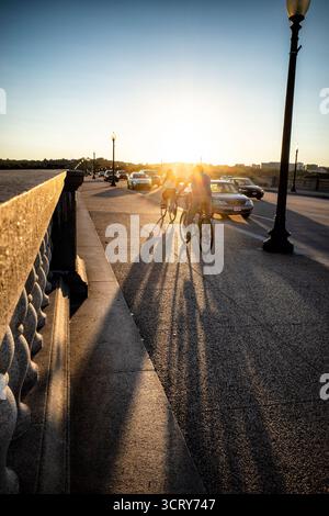 Arlington Memorial Bridge At Sunset Washington DC | WASHINGTON D.C., United States — Two cyclists ride the sidewalk of Arlington Memorial Bridge into a low sunset, their long shadows stretching across the granite deck as traffic streams past. The bridge, built 1926-1932 to a design by McKim, Mead and White, links the Lincoln Memorial to Arlington National Cemetery along a sightline chosen to symbolize post-Civil War reunification. Its granite balustrades and lampposts frame a roughly 2,138-foot (652 m) crossing of the Potomac River. Stock Photo
