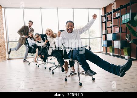 Energetic team having fun racing office chairs in a bright modern workplace, showcasing teamwork Stock Photo