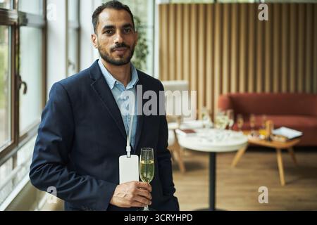 Portrait of young adult Middle Eastern man standing indoors holding champagne glass and wearing conference badge, looking at camera with confident expression, modern lounge background visible Stock Photo