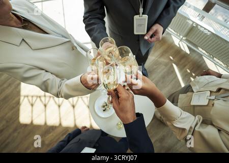 Group of middle aged multiethnic professionals clinking champagne glasses during business celebration, standing around table with snacks, wearing formal attire and conference badges Stock Photo