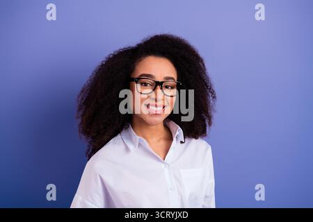 Positive young woman with glasses wearing a white shirt standing against a purple background and smiling warmly Stock Photo