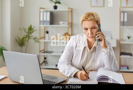 Focused mature woman working at desk with laptop, taking notes while making work calls on phone. Stock Photo