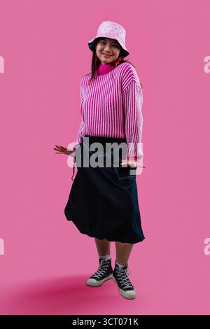 Playful asian woman in a pink patterned bucket hat and striped chunky sweater, smiling and pointing at camera against a vibrant pink studio backdrop, Stock Photo