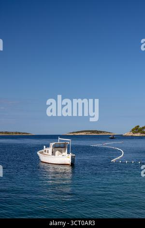 A serene scene of a white boat floating gently on calm blue water, with a curved line of buoys and distant islands under a bright, clear sky. Stock Photo