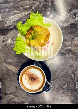 A frothy cappuccino with cinnamon served in a blue cup alongside kaya toast on a marble table with a salad and bread dish in background ipoh, Stock Photo