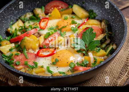 Fried eggs and potatoes, tomatoes, zucchini  and chili peppers on wooden background. Stock Photo