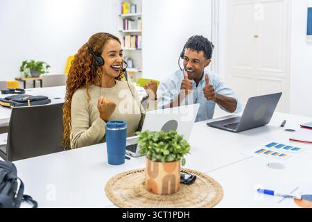Two diverse call center agents wearing headsets and working on laptops, celebrating their achievement with joy, showing thumbs up and a triumphant fis Stock Photo