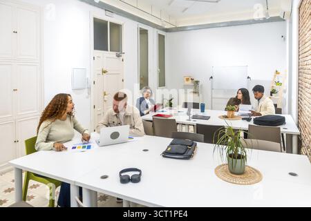 Group of diverse business professionals discussing ideas, analyzing documents, and working on laptops during a collaborative meeting in a bright, mode Stock Photo