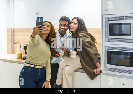 Diverse coworkers cheerfully taking a selfie on a smartphone during a relaxed office kitchen break, sharing laughs and casual bonding over coffee and Stock Photo
