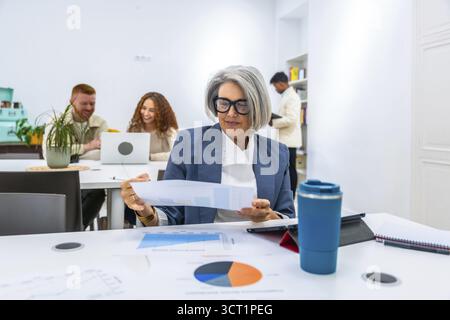Mature female manager reviewing a financial report with charts and graphs on a table, collaborating with diverse young coworkers in a modern office wo Stock Photo