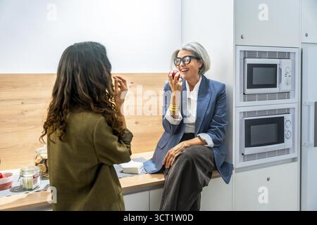 Two business women colleagues having a healthy snack, eating apples and laughing together while taking a break in the modern office kitchen or canteen Stock Photo