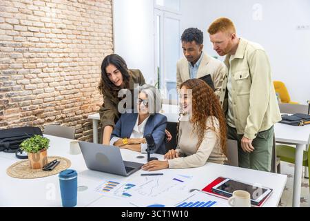 Diverse business professionals working together around a laptop, analyzing financial charts and presenting ideas during a casual office meeting, showc Stock Photo