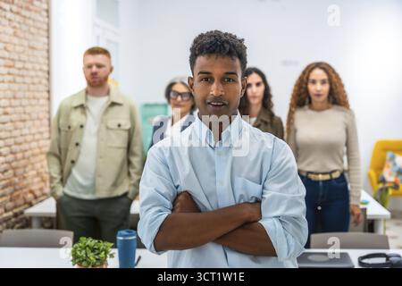 Young man standing with arms crossed, confidently looking at the camera, with his diverse team members blurred in the background, working together in Stock Photo