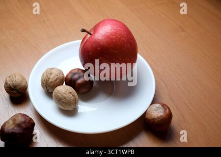 A bright red apple sits on a white plate surrounded by various nuts on wood. Stock Photo