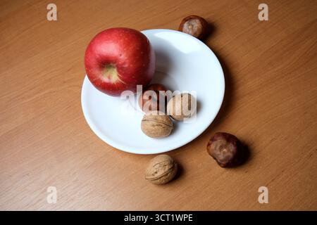 A shiny red apple sits with different nuts on a white plate atop a warm wood surface. Stock Photo
