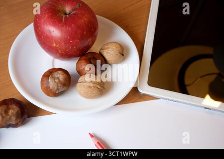 A red apple sits beside mixed nuts on a white plate next to a tablet and paper. Stock Photo