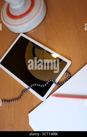 A bright tablet lies on a wooden desk beside a red pencil and white papers. Stock Photo