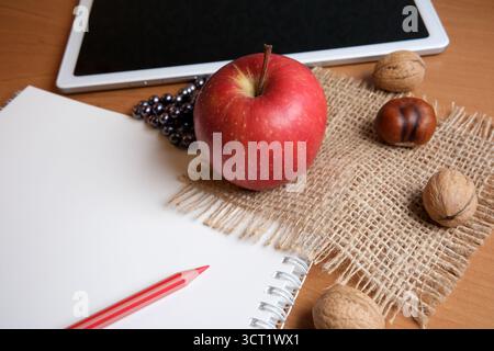 A red apple sits on a burlap cloth next to a blank sketchbook and tablet, with nuts. Stock Photo