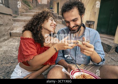 A happy young couple enjoys a romantic date, eating fresh pasta and toasting with white wine on the stone steps of a historic Italian alley. Stock Photo