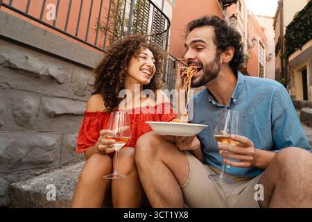 A playful young woman laughs as she feeds her smiling partner a forkful of spaghetti during a romantic lunch date on the steps of a historic Italian a Stock Photo
