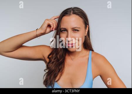 A woman with long, wavy hair poses confidently while touching her head with a playful expression. She wears a light blue top that highlights her cheer Stock Photo