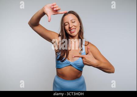 A young woman wearing blue activewear poses joyfully in a studio. She smiles widely while framing her face with her hands, expressing happiness and pl Stock Photo