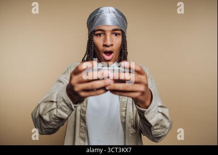 A young man with braided hair and a silver head wrap holds a gaming console, revealing a surprised expression as he reacts to a gaming moment. The ind Stock Photo