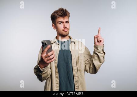 A young man stands indoors, holding a smartphone in one hand and pointing upward with the other. He looks thoughtful and engaged in a conversation, su Stock Photo