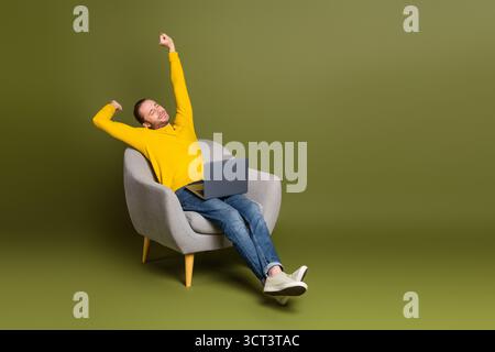 Young man in yellow sweater stretching with laptop in stylish chair against green background Stock Photo