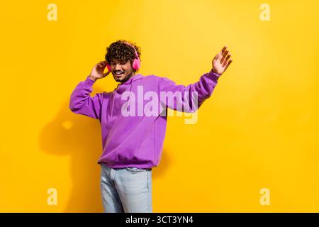 Cheerful young man in a purple hoodie enjoying music through headphones against a vibrant yellow background Stock Photo