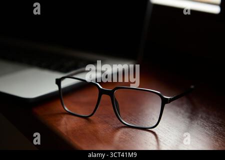 Pair of eyeglasses placed on a polished wooden desk next to an open laptop, creating a stylish workspace still life scene with professional atmosphere Stock Photo