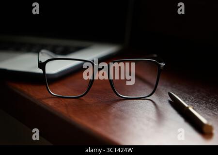 Black eyeglasses resting on a polished wooden desk alongside a pen with a blurred laptop background. Minimal workspace still life for office and techn Stock Photo