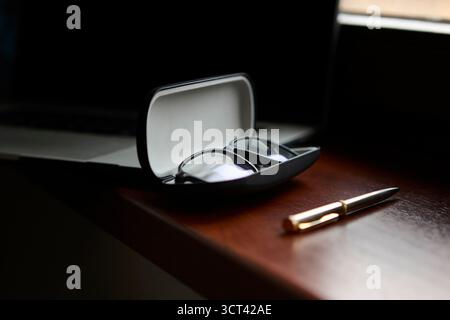 Open glasses case with eyeglasses inside resting on polished wooden desk beside pen and blurred computer in background. Clean product still life. Stock Photo
