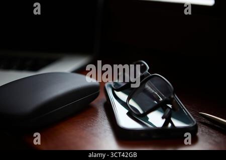 A folded pair of eyeglasses lies on a polished wooden desk next to a protective glasses case and a pen. The scene suggests an organized office or home Stock Photo