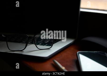 Eyeglasses are positioned on a laptop keyboard with a pen and smartphone placed on the desk beside it. The setting conveys a modern business or academ Stock Photo
