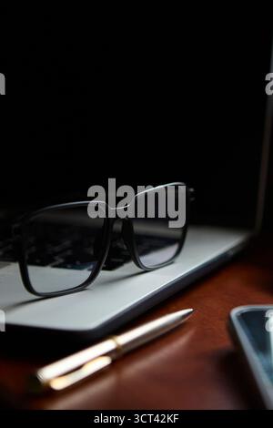 A pair of eyeglasses and a golden pen are resting on a polished wooden desk in front of a laptop. The scene suggests a thoughtful working or studying Stock Photo