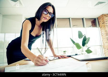 Confident young businesswoman working in an office using documents and laptop, symbolizing professionalism and success Stock Photo