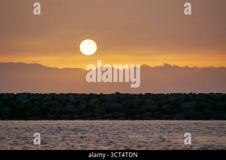 A serene sunset over a calm sea, with a large sun setting behind distant mountains. The sky is painted in warm hues of orange and yellow. Stock Photo