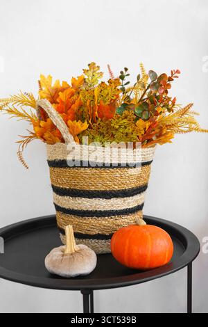 Woven basket with colorful autumn flowers and yellow leaves on black table with small pumpkin on white wall background, creates fall atmosphere of liv Stock Photo