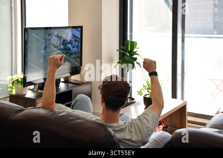 Young man sitting on a couch at home watching American football on television with raised arms celebrating a play Stock Photo