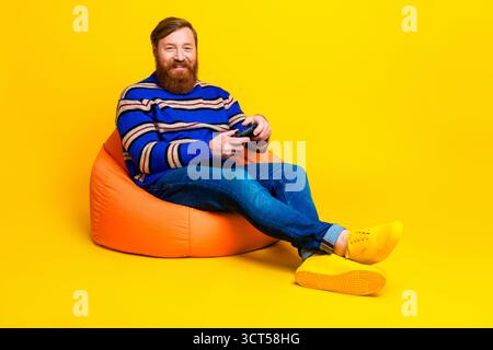 Trendy man with red beard seated on orange beanbag and playing video game against bright yellow background Stock Photo