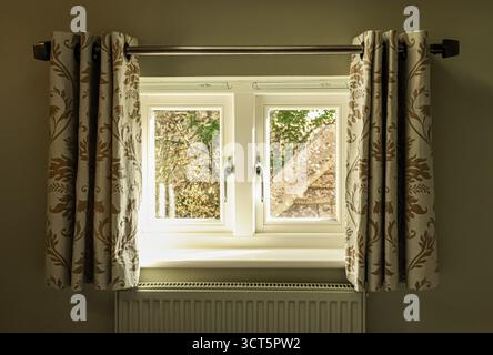 Window with patterned curtains and radiator, letting in warm sunlight and creating a cozy home interior. Cotswolds countryside, England Stock Photo