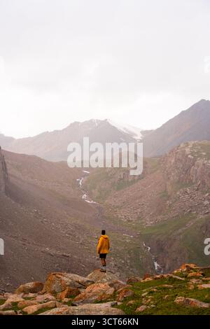 A male hiker (20s-30s) in a bright yellow jacket stands on a boulder overlooking a vast, rugged mountain valley with a winding stream below Stock Photo