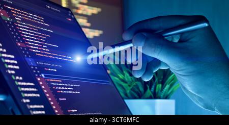 Close-up of a hand holding a stylus, interacting with source code on a screen in a modern programming environment with blue lighting. Stock Photo