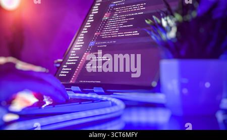 Close-up of a developer typing on a keyboard in a vibrant workspace with code on screen and glowing blue-purple lighting. Stock Photo