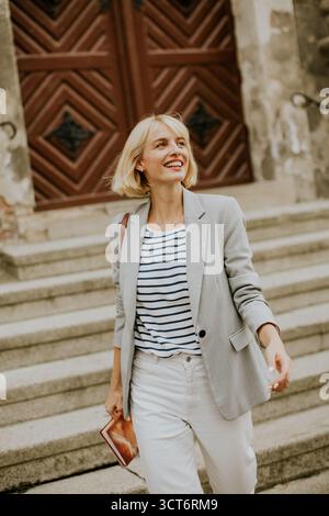 A young woman wearing a light blazer and striped shirt walks down stone steps, smiling happily. The background features large wooden doors and a vinta Stock Photo
