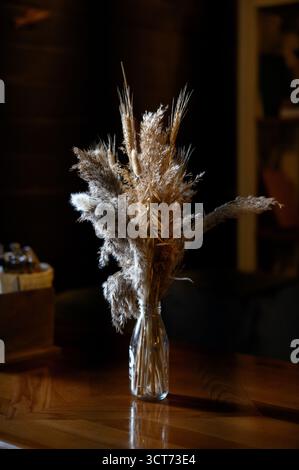 Still life with fluffy dried flowers in a bottle in a cafe interior with subdued lighting. Decorative composition of grain ears in a transparent vase Stock Photo