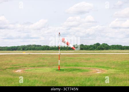 Red and white windsock near runway in a grassy field under a cloudy sky with trees in the background. Symbol of wind direction and speed, commonly use Stock Photo